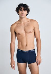 Boxer-briefs bleu marine avec une ceinture de marque blanche, design ajusté, en coton, et texture lisse. Le modèle se tient debout sur un fond clair.