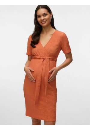 Femme souriante, vêtue d'une robe portefeuille orange texturée, longueur genou, à manches courtes, tenant son ventre, debout devant un fond uni.