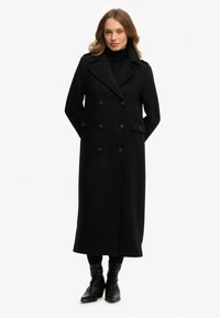 Lange zwarte wollen jas met een dubbelrijig ontwerp, grote knopen en zijzakken. Gestyled over een zwarte coltrui en gedragen met enkellaarzen.