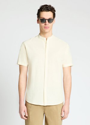 Mann trägt cremefarbenes kurzärmeliges Hemd mit Mandarin-Kragen, beige Hose und dunkle Sonnenbrille, steht vor einfarbigem Hintergrund.