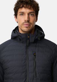 Donkergrijze pufferjack met rits aan de voorkant, verstelbare capuchon en borstzak. Voorzien van horizontale stiknaden en een gladde afwerking.