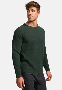 Pull en maille côtelée verte avec un col rond et des manches longues, associé à un jean foncé. Tissu texturé et coupe ajustée.