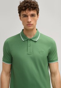 Grüne Polohemd mit klassischem Kragen, kurzen Ärmeln und kontrastierendem weißen Piping am Kragen und den Ärmeln. Gesticktes Logo auf der Brust.