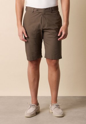 Uomo che indossa pantaloncini marroni fino al ginocchio, camicia bianca infilata e scarpe beige chiare con lacci, in piedi su un pavimento liscio e neutro.