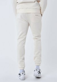 Pantaloni della tuta color crema in morbido materiale, con polsini elasticizzati e dettaglio del logo sul retro. Indossati con sneakers bianche e calzini blu.