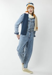 Jeansoverall met lange mouwen, blauwe kleur en een lichte wassing, gecombineerd met een gestreepte gebreide muts en sjaal in oranje, blauw en crème.