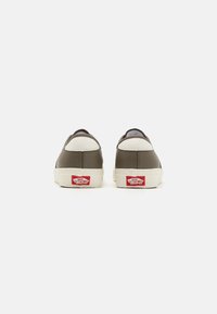 Zapatillas de cuero marrón con acentos crema, punta redondeada, suela blanca plana y detalle del logo rojo en el talón. Textura suave en la superficie superior.