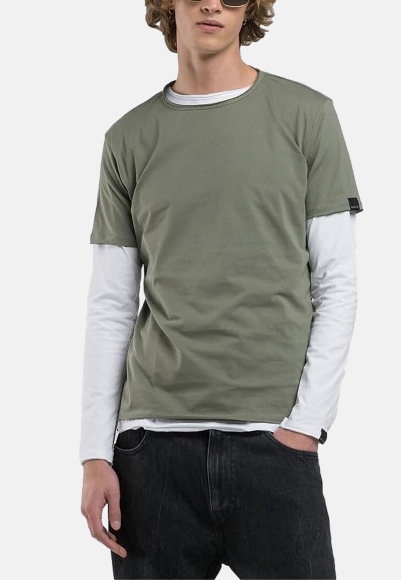 T-shirt olive green à manches courtes superposé sur une chemise blanche à manches longues, avec un col rond et une finition texturée, associé à un jean foncé.