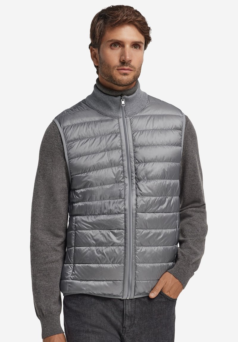 Szara ocieplana kamizelka z wysokim kołnierzem, zapinana na zamek błyskawiczny, z poziomym pikowaniem, noszona na ciemnoszarej koszuli z długim rękawem.