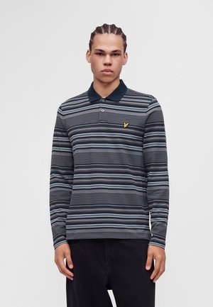 Jeune homme portant un polo à manches longues rayé avec un col bleu marine et un logo d'oiseau jaune sur un fond uni.