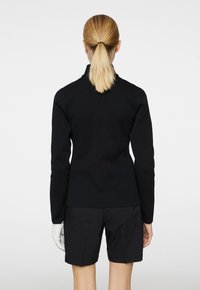 Svart stickad tröja med hög krage, texturerad mönster och långa ärmar, kombinerad med svarta shorts och en vit handske på ena handen.