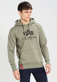 Olivgrüner Hoodie aus weicher Baumwolle mit einer Vordertasche. Verfügt über ein schwarzes Logo und ein rotes Stoffetikett am Saum für Akzente. Verstellbare Kapuze.