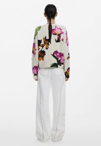 Pull à motif floral crème avec des fleurs roses, violettes et oranges ; poignets et ourlet côtelés. Associé à un pantalon blanc à jambes larges avec des coutures contrastantes.