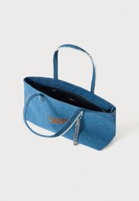 Džínová taška na rameno s dvoma rukoväťami, modrej farby, s oranžovým logom, čiernym vnútrom a retiazkovým doplnkom na jednej strane.