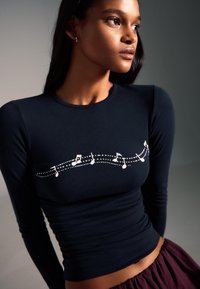 Fekete, hosszú ujjú ing, sima anyagból, fehér kottafej dizájnnal és pöttyös vonalakkal a mellrészen.