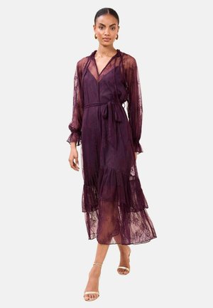 Žena u prozirnoj ljubičastoj midi haljini od čipke s dugim rukavima, remenom u struku i naboranim rubom, uparena s bež sandalama na visoku petu.