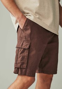 Braune Cargo-Shorts aus strukturiertem Stoff, mit seitlichen Taschen und lockerer Passform. Tragen mit einem hellen Hemd.