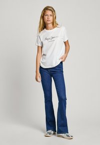 Vit bomullströja med skriptlogotypen "Pepe Jeans London" framtill, ihop med mörkblå utsvängda jeans och silverfärgade sneakers.