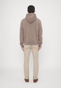 Felpa marrone con vestibilità rilassata, polsini e cintura a coste; indossata con pantaloni beige e scarpe marroni, vista da dietro.