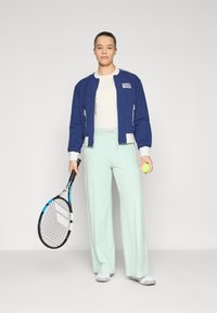 Casaco de bombardeiro azul-marinho com detalhes brancos, calças largas menta claras, segurando uma raquete de ténis e uma bola, com fundo neutro.