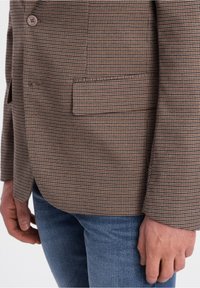 Brązowa marynarka w kratę z teksturowanej tkaniny, zapinana na jeden guzik, z klapami z wycięciami. W zestawie z niebieskimi dżinsami.