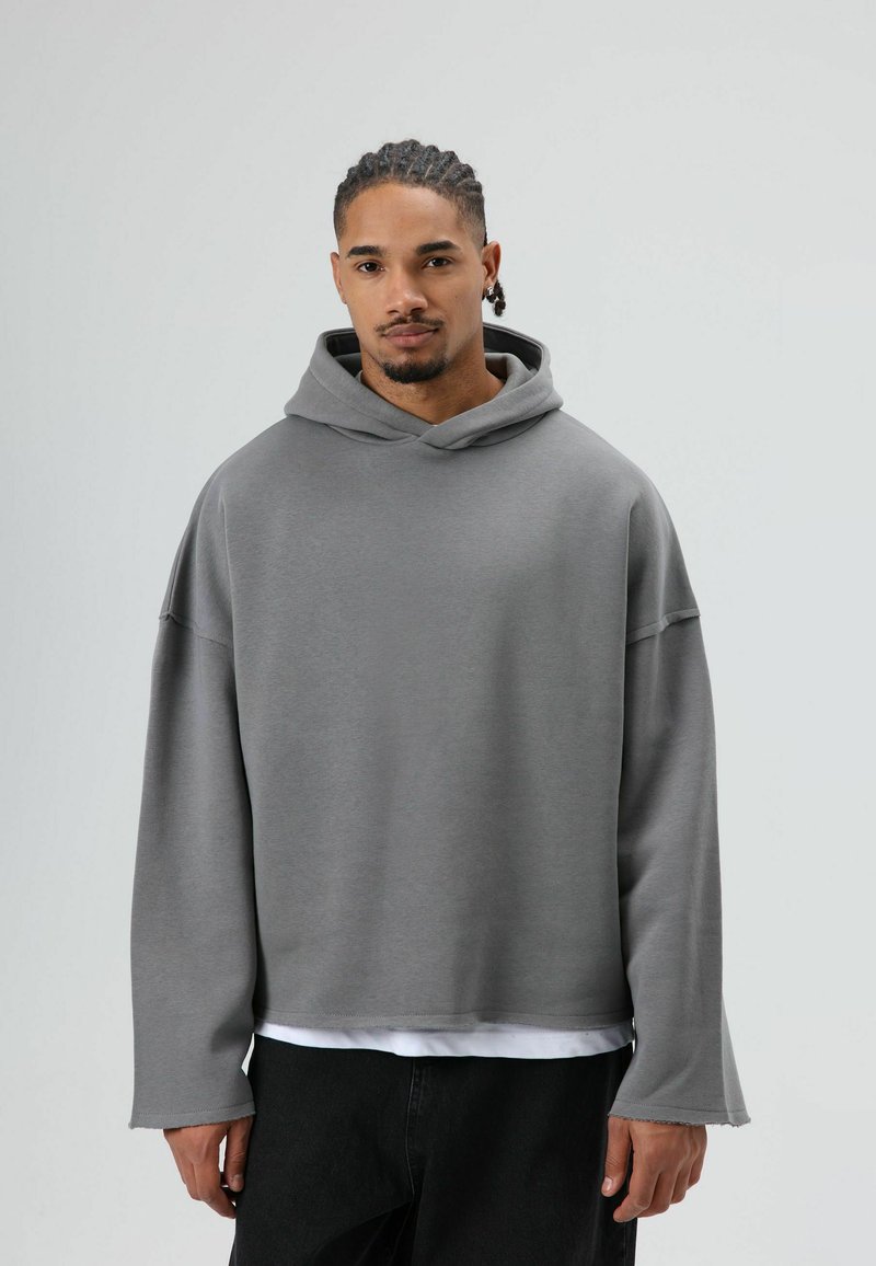 Grijze oversized hoodie met trekkoord aan de capuchon, lange mouwen en een zachte textuur, gecombineerd met een wit shirt eronder en donkere broek.