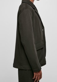 Person klädd i en mörk olivgrön blazer med fickor med lock, knäppt framtill och matchande byxor, sedd från sidan mot en enfärgad bakgrund.