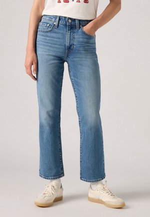 Person trägt mittelblaue gerade Jeans, weiße Socken, cremefarbene Sneakers und ein weißes Shirt mit Grafikdruck, Hand in der Tasche.