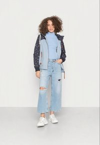 Casaco azul claro com mangas de padrão de estrelas em azul marinho, combinado com jeans azuis desbotados e rasgados e ténis brancos. Superfície texturizada e cores contrastantes.