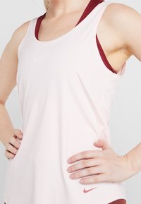 Top da allenamento rosa chiaro realizzato in tessuto morbido, con un design racerback bordeaux e un piccolo logo Nike rosso sul basso davanti.