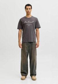 Mørkegrå bomulds T-shirt med grafisk print på forsiden, parret med løse olivengrønne denimbukser og beige fodtøj.