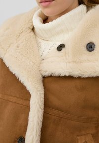 Bruine suède jas met een crèmekleurige namaakbont kraag, voorzien van twee zwarte knopen en een getextureerde witte gebreide coltrui eronder.