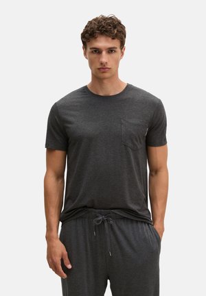 Młody mężczyzna z kręconymi włosami, ubrany w ciemnoszary t-shirt z krótkim rękawem i pasujące spodnie ze ściągaczem, stoi na jednolitym tle.