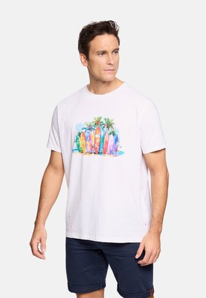 Hombre con camiseta blanca con diseño colorido de tablas de surf y palmeras, combinado con pantalones cortos oscuros remangados, de pie frente a un fondo liso.