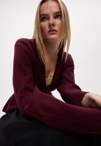 Bordový cardigan s hlbokým výstrihom do V a dlhými, rozšírenými rukávmi. Mäkká textúra, zapínanie na gombíky vpredu, nosený cez svetlo béžový top.