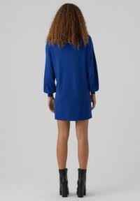 Knälång blå klänning med långa puffärmar, gjord av mjukt tyg. Synliga sömmar vid fållen, tillsammans med svarta klackstövlar med tjock klack.