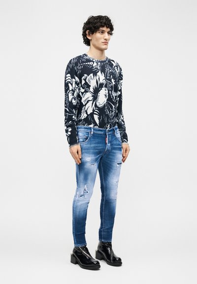 Άνδρας στέκεται φορώντας μαύρο και λευκό πουλόβερ με λουλούδια, σκισμένο στενό τζιν μπλε και μαύρα μπότακια με τακούνι σε λευκό φόντο.
