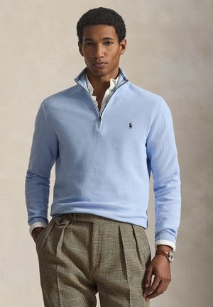 Mann trägt hellblauen Viertel-Reißverschluss-Pullover über weißem Hemd und brauner karierter plissierter Hose, steht mit einer Hand in der Tasche.