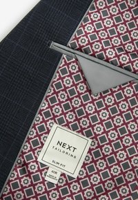 Intérieur d'une veste en tissu texturé bleu marine avec un motif géométrique en bordeaux, gris et crème. L'étiquette indique "NEXT TAILORING SLIM FIT 40R."