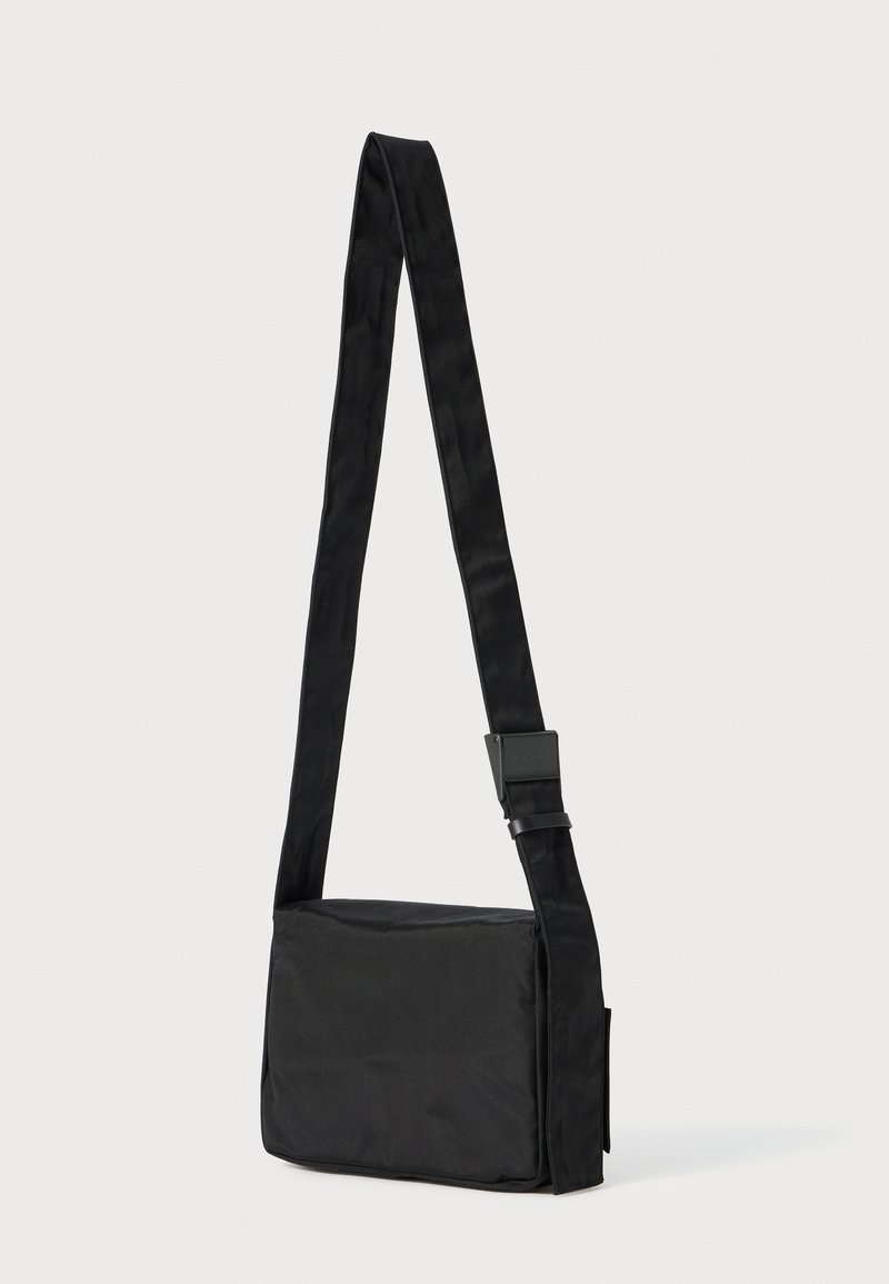 Sort skuldertaske lavet af glat stof med rektangulær form, justerbar rem og enkelt spænde til sikker bæring.