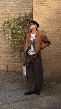 Brown zip-up jacket over a graphic t-shirt, wide brown pants, and dark shoes. Accessories include sunglasses and a black hat. Brick wall background.