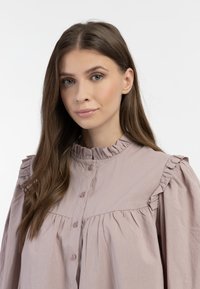 Jasnoróżowa bluzka uszyta z miękkiego materiału, z falbaną przy dekolcie i bufiastymi rękawami, z zapięciem na guziki z przodu.