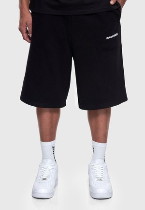 Pantaloni scurți negri până la genunchi cu logo-ul „Dropsize”, purtați cu șosete albe și adidași albi.