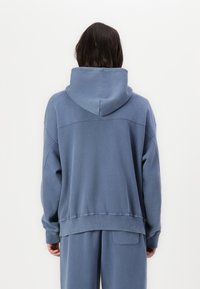 Sweat à capuche bleu atténué, offrant une coupe décontractée, des épaules tombantes et des poignets côtelés. Texture douce avec un détail de couture au dos.