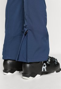 Marineblå skibukser med en glat tekstur, sidelukninger ved stropperne, båret med sorte skistøvler med hvide accenter og formet hæl.