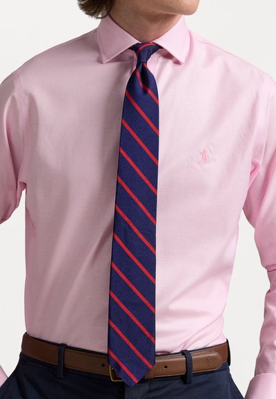 Ανοιχτό ροζ πουκάμισο με γιακά, συνδυασμένο με ναυτικό μπλε γραβάτα με κόκκινες διαγώνιες ρίγες. Καφέ ζώνη που αντιπαραβάλλεται με σκούρα παντελόνια.