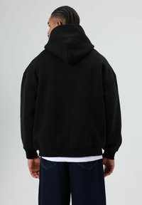 Zwarte capuchontrui met afgezakte schouders, geribde zoom en manchetten, gedragen over een wit T-shirt, gezien vanaf de achterkant.