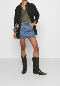 Persona che indossa una camicia oversize nera, un top verde oliva, una minigonna di jeans, stivali cowboy neri e una borsa a tracolla con stampa leopardata.