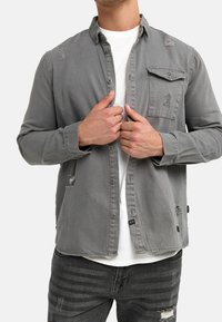 Grijze denim blouse met knopen en een zak, voorzien van versleten accenten op de schouders en zoom. Draagt over een wit T-shirt.