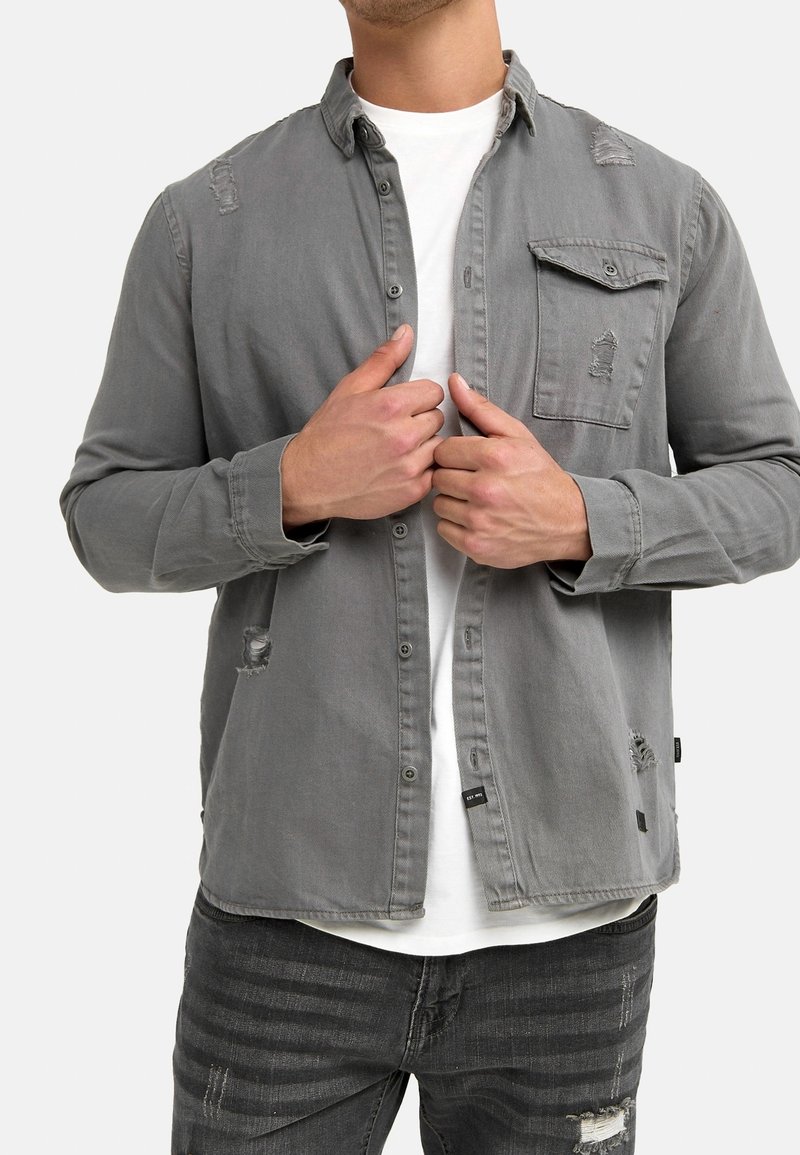 Grijze denim blouse met knopen en een zak, voorzien van versleten accenten op de schouders en zoom. Draagt over een wit T-shirt.